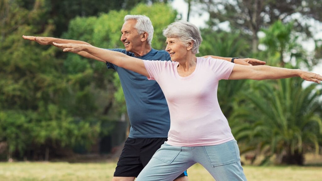 Stretching e declino cognitivo: una donna e un uomo anziani praticano la posizione del guerriero yoga al parco.