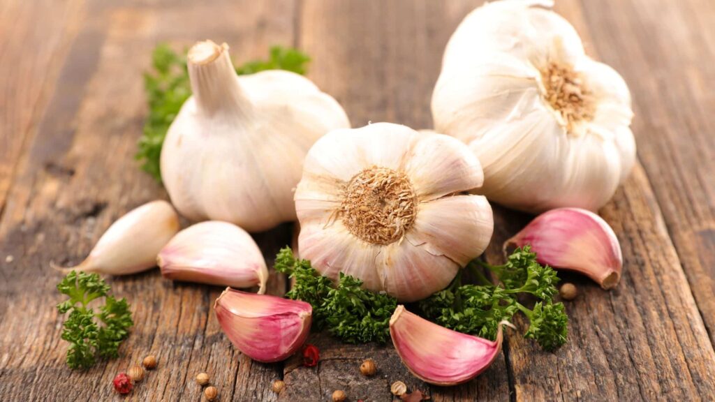 Tre agli interi e 3 spicchi d'aglio su una tavola di legno con alcune erbette - benefici aglio crudo e allicina